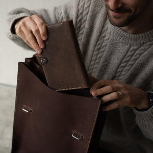 Man holding a brown leather bag while putting a brown leather journal into the bag. 